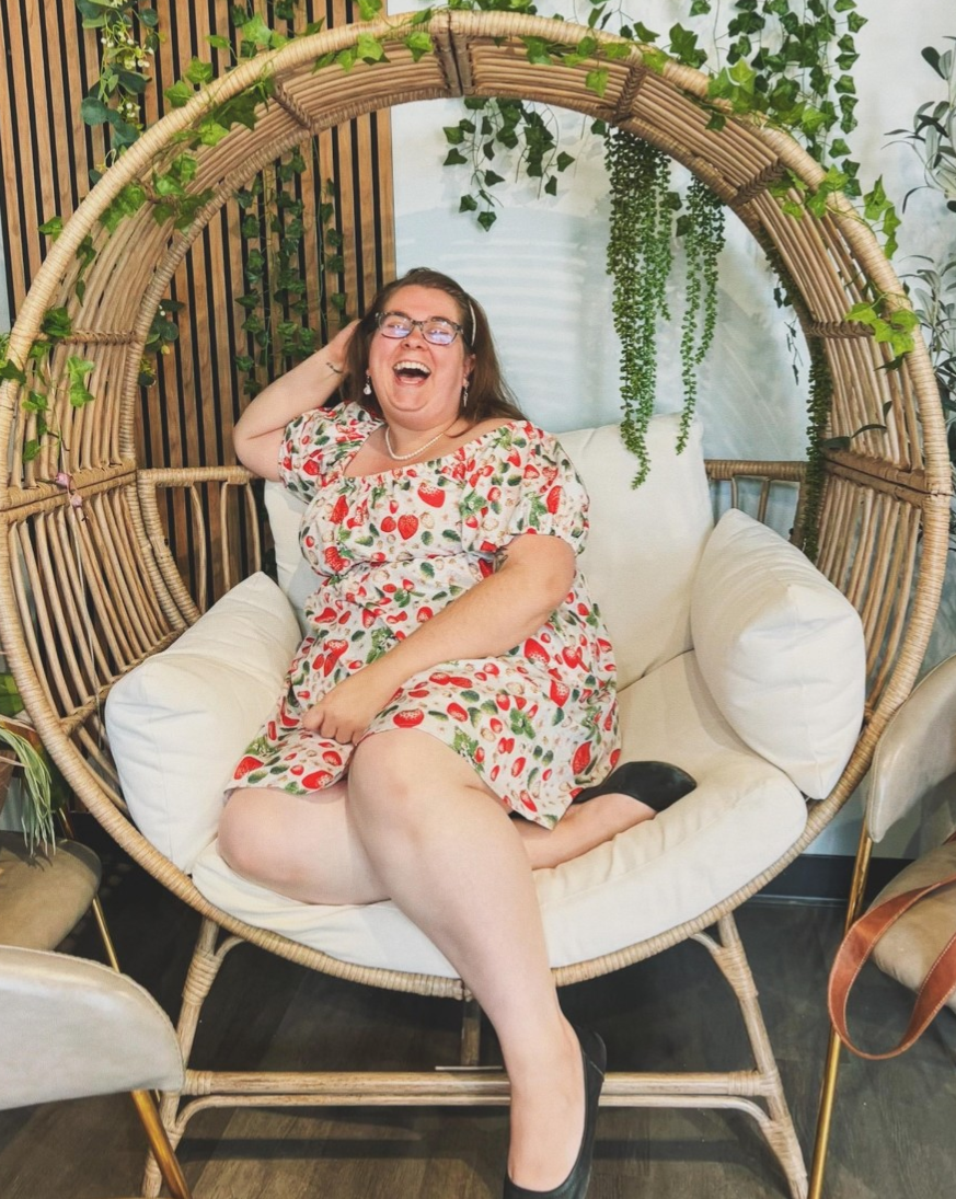 A person sitting in a wicker chair, wearing a floral dress with a pattern of strawberries, laughing and looking cheerful.