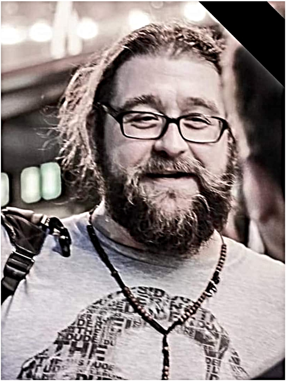 A smiling man with a beard and glasses, wearing a t-shirt and a beaded necklace.