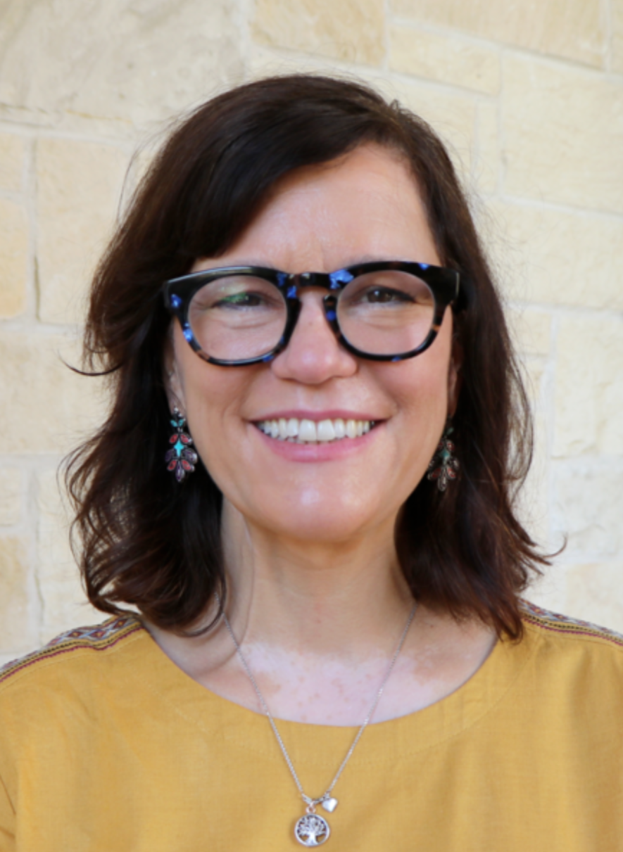  A woman with shoulder-length brown hair, tortoiseshell glasses, and drop earrings smiles at the camera against a neutral background. 