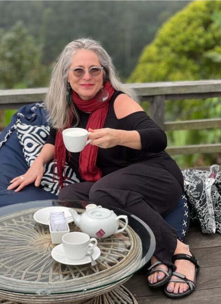 A woman with gray hair, sunglasses, and a red scarf sits outdoors holding a white teacup, with a tea set on the table in front of her.