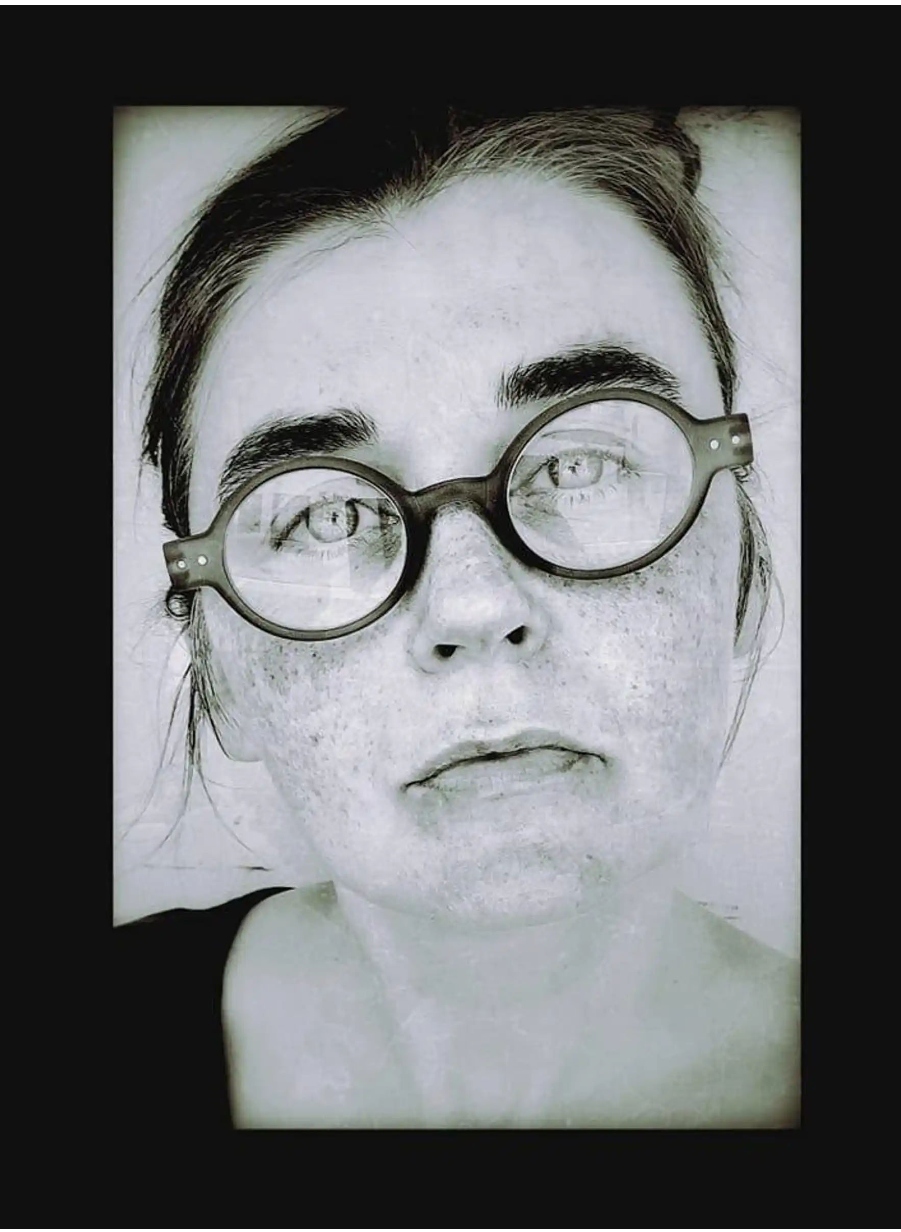 A close-up, black-and-white portrait of a person with glasses, looking directly at the camera. They have freckles and a serious expression.