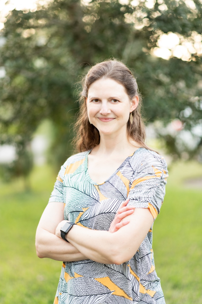 A woman with long hair, wearing a patterned shirt, stands outdoors with her arms crossed and a slight smile.